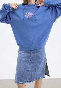 Sudadera con capucha en azul, con un gráfico rosa de impresión "San Diego". Acompañada de una falda de mezclilla azul claro con una abertura lateral.