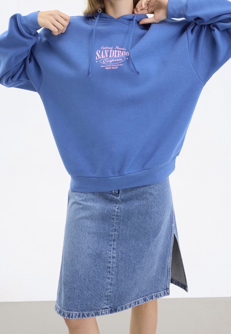 Sudadera con capucha en azul, con un gráfico rosa de impresión "San Diego". Acompañada de una falda de mezclilla azul claro con una abertura lateral.