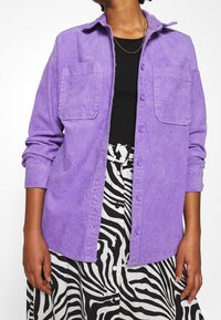 Purple corduroy button-up shirt with two chest pockets, long sleeves, and a relaxed fit, worn over a black top and zebra-print skirt.