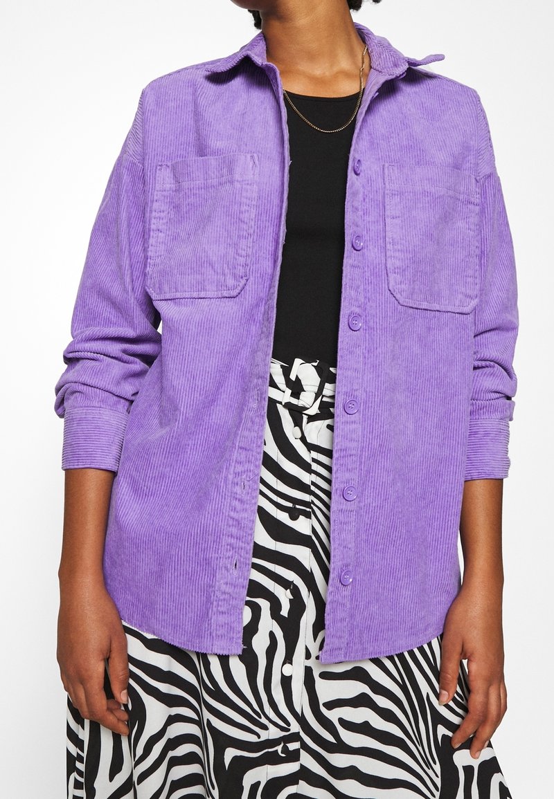 Purple corduroy button-up shirt with two chest pockets, long sleeves, and a relaxed fit, worn over a black top and zebra-print skirt.