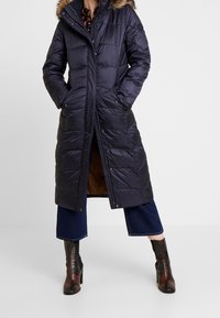 Tmavě modrý prošívaný kabát s odepínací kapucí s kožešinovou podšívkou, zapínáním na knoflíky, bočními kapsami a prošitým vzorem.