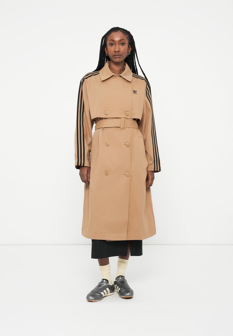 Beige trench coat med dobbeltknaplukning, oversized design og sorte striber. Stylet med grå sneakers og hvide sokker.