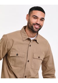 Man met kort donker haar en baard die glimlacht, gekleed in een beige overhemd met knopen en twee borstzakken tegen een witte achtergrond.