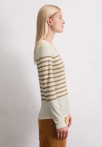 Vrouw met steil blond haar draagt een crèmekleurige bovenkant met lange mouwen met horizontale bruine strepen en een bruine rok, staande tegen een witte muur.