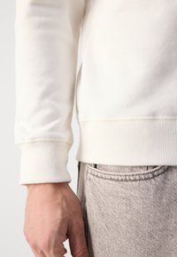 White sweatshirt with ribbed cuffs, paired with light gray denim pants featuring a subtle diagonal pattern. Close-up of sleeve and hand.