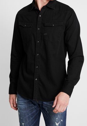 Homme portant une chemise noire à manches longues avec deux poches poitrine et un jean bleu usé, debout devant un fond uni.