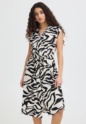 Kobieta z długimi kręconymi włosami, ubrana w czarno-białą sukienkę w wzór zebry bez rękawów z paskiem w talii, stojąca na jasnym jednolitym tle.