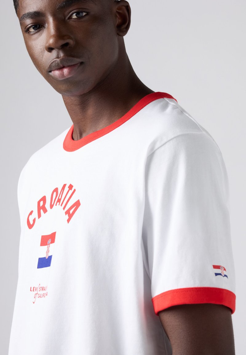 Young man wearing a white T-shirt with red trim, "CROATIA" text, a flag graphic on the chest and sleeve, standing against a plain background.