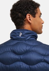 Navy blue puffer jacket with a quilted texture, high collar, and a small logo on the back. Smooth fabric with a sleek finish.