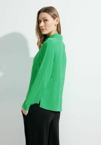 Blusa verde trasparente con colletto, maniche lunghe e spacchi laterali, abbinata a pantaloni neri. Tessuto leggero con una consistenza liscia.
