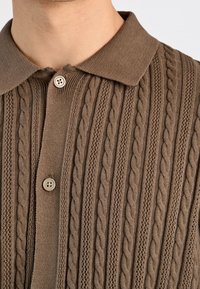 Ruskea neulottu polo-paita, jossa on kaapeliteksti, kahdella vaalean beige-napilla, henkilö, jonka pää näkyy kaulasta rintaan.