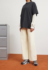 Oversized donkergrijze T-shirt gelaagd over een witte longsleeve, gecombineerd met crèmekleurige corduroy broek met wijde pijpen en grijze sportschoenen.