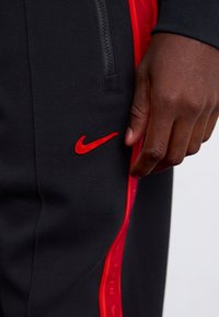 Črne športne hlače z rdečim logotipom Nike in rdečimi stranskimi poudarki. Material ima teksturirano površino, z žepom na zadrgo za shranjevanje.