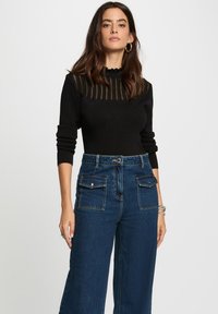 Femme avec de longs cheveux bruns portant un haut noir à manches longues avec des rayures transparentes et un jean large taille haute bleu, se tenant contre un fond uni.