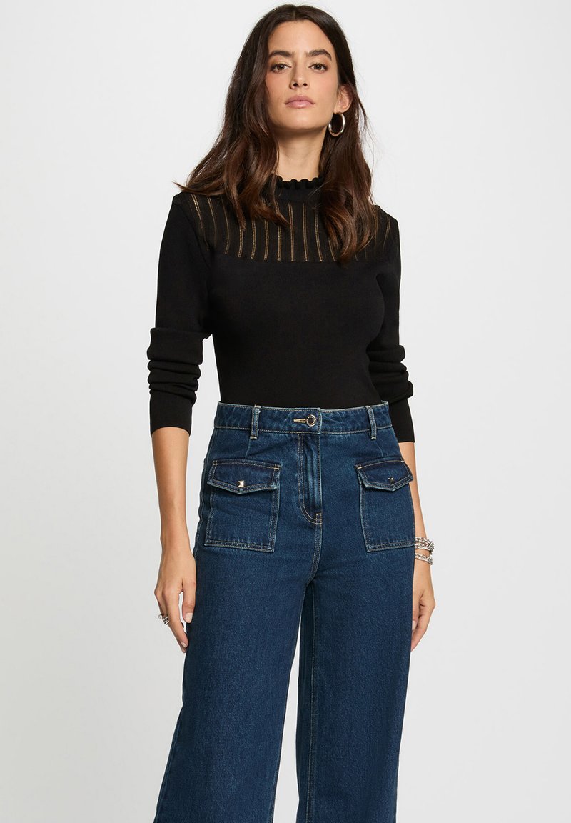 Femme avec de longs cheveux bruns portant un haut noir à manches longues avec des rayures transparentes et un jean large taille haute bleu, se tenant contre un fond uni.
