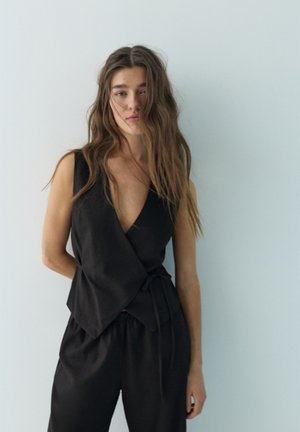 Jonge vrouw met lang los haar draagt een mouwloze zwarte overslagtop en bijpassende zwarte broek, staand tegen een effen lichtblauwe muur.