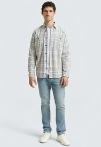 Chemise à carreaux beige et bleue à boutons, avec deux poches poitrine, portée sur un t-shirt blanc, associée à un jean bleu clair et des baskets blanches.