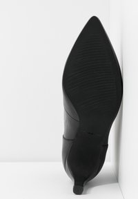 Zapato de tacón alto negro con punta afilada, suela de goma texturizada, parte superior de cuero suave y diseño minimalista, que incluye un tobillo acolchado.
