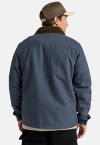 Dark blue jacket with a brown, textured collar, featuring a snap closure and long sleeves. Worn over a gray, ribbed sweater.