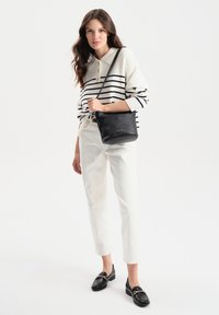 Czarna skórzana torba na ramię z teksturowanym wykończeniem, noszona przez osobę w paskowanym swetrze w kolorze kremowym i granatowym oraz białych spodniach, w połączeniu z czarnymi loafersami.