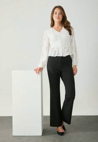 Blusa blanca texturizada con bajo peplum y botones, combinada con pantalones negros acampanados. La modelo está de pie junto a un pedestal blanco, luciendo tacones.