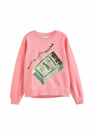 Pink sweatshirt med en grafik af en grøn matcha-tepakke og teksten "We are the perfect matcha" hældt over den.
