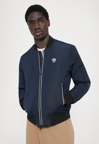 Casaco bomber azul-marinho com fecho frontal, punhos canelados e um pequeno logotipo no peito. Feito de um material suave e leve.