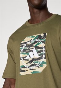 Mand med olivengrøn T-shirt med camouflage-musteret Adidas-logo og teksten "Mærket med de tre striber" på brystet.