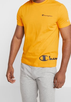 Hombre que lleva una camiseta Champion de color naranja brillante y pantalones deportivos gris claro, de pie con los brazos relajados a los lados.