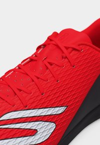 Κόκκινο αθλητικό παπούτσι με υφή στην επάνω επιφάνεια, κόκκινες κορδόνια, μικρές οπές αερισμού και μεγάλο λευκό και μαύρο λογότυπο στο πλάι.