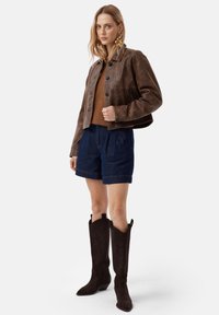Veste en cuir brun raccourcie sur un pull brun, associée à un short en denim bleu marine et à de longues bottes en daim marron foncé à bouts pointus.