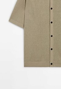 Cardigan en maille beige à manches courtes avec des boutons noirs sur le devant, poignets et ourlet côtelés, présenté à plat sur fond blanc.