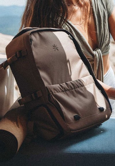 Mochila de tela marrón con correas ajustables, bolsillo frontal y detalle del logo. Incluye una esterilla enrollada al lado sobre una superficie azul.