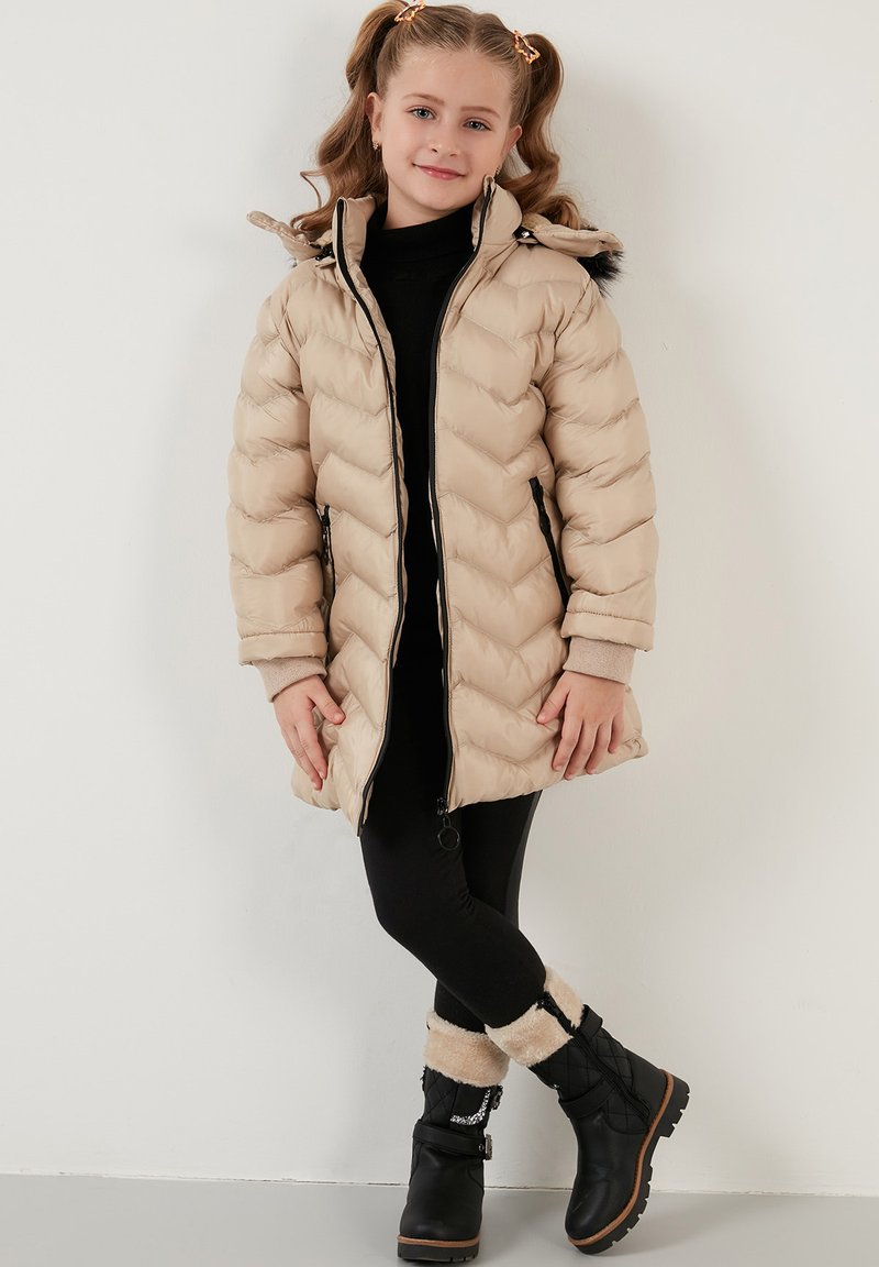 Beige quiltet puffjakke med hætte, med sort lynlås lukning og sidelommer. Båret med sorte leggings og fuzzy-boots accenter.