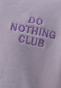 Vaalean lilaa kangasta, jossa on tummalla lilalla kirjailtu teksti "DO NOTHING CLUB", ja jossa on sileä, hieman teksturoitu pinta.