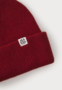 Knitted beanie in deep red with ribbed texture. Features a white label with a circular black logo on the fold. Soft, stretchable material.