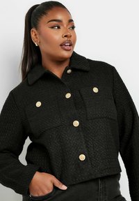Svart kort jacka i texturerat tyg med krage, två bröstfickor och guldfärgade knappar som hårdvaruaccent.