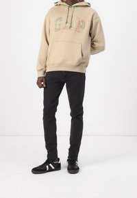 Beżowa bluza z kapturem z logo "GAP" w wielokolorze, kieszenią kangurką, z warstwowym kapturem w kratę. Połączona z czarnymi, dopasowanymi jeansami i czarnymi trampkami.