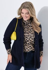 Mörkblå stickad cardigan med knäppning, bärs över en leopardmönstrad topp. Accessoarer inkluderar en liten gul väska. Mörka jeans syns.
