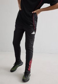 Melna sporta apģērba komplekts, kas sastāv no īsu piedurkņu topa un šaurām biksēm ar sarkanām malām. Apavi ir melnā un zaļā krāsā.