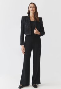 Schwarze kurze Blazer mit strukturiertem Stoff, breiten Revers und Knopfverschluss, kombiniert mit langen schwarzen Hosen und flachen schwarzen Schuhen.