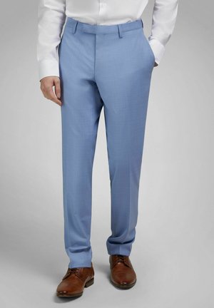 Mann trägt hellblaue Anzughose, weißes Hemd mit Knöpfen und braune Lederschuhe, steht mit einer Hand in der Tasche.