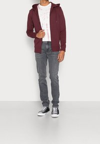 Hombre con una sudadera burdeos con cremallera, camiseta de tie-dye blanca y rosa, jeans grises y zapatillas altas negras de pie contra una pared blanca.