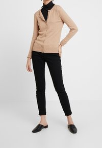 Beige kofta med rund halsringning, knappstängning och ribbad fåll. Kombinerad med svarta smala byxor och svarta slip-on skor.