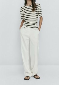 Randig svart och beige t-shirt med avslappnad passform. Vita vida byxor av texturerat tyg. Svarta flipflops. Enkel, modern design.