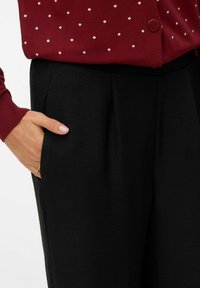 Personne portant un pantalon noir avec une main dans la poche et un pull rouge à pois blancs avec un bouton à la taille.
