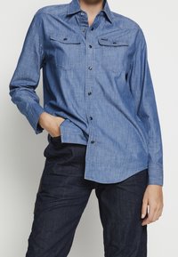 Person i blå denimskjorta med knappar och fickor samt mörkblå jeans, med en hand i fickan, mot en enfärgad bakgrund.