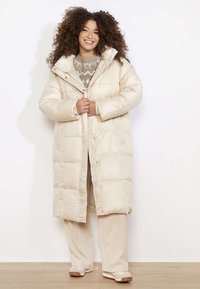 Długa, beżowa kurtka puchowa z kapturem, zapinana na zamek i zatrzaski. Posiada pikowany wzór i szerokie rękawy, noszona na wzorzysty sweter.