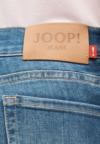 Κοντινή λήψη μπλε τζιν με δερμάτινο ταμπελάκι χρώματος ταμπά με την ένδειξη "JOOP! JEANS" και μικρή κόκκινη υφασμάτινη ετικέτα με θαυμαστικό.