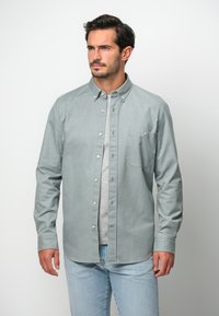 Camicia button-up verde chiaro in cotone, con taschino frontale, colletto e maniche lunghe, abbinata a jeans blu chiaro.